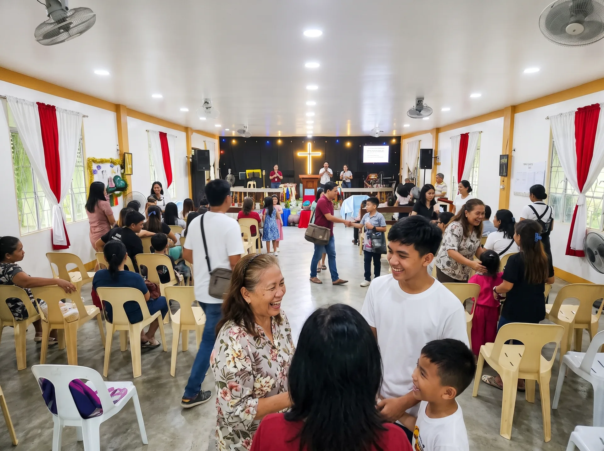 Smiling church members talking together in the aisle after a service at PCVCCI, with monobloc seating and the sanctuary cross visible in the background